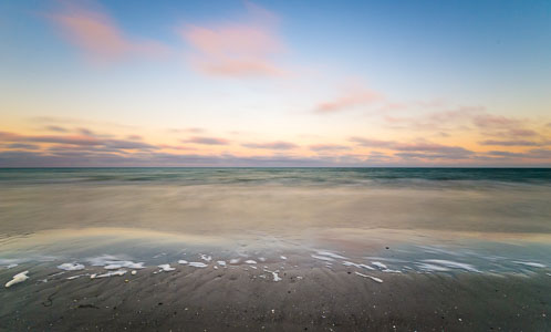 Ostseeufer und Meerschaum am Darß – Die Aufnahme zeigt den Flachwasserbereich vor Prerow nach Sonnenuntergang. Der Himmel über dem Horizont leuchtet in kräftigen Gelb- und Orangetönen.