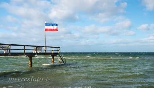 Eine kleine Seebrücke am Strand von Niendorf mit einer Schleswig-Holstein-Flagge im Sturm