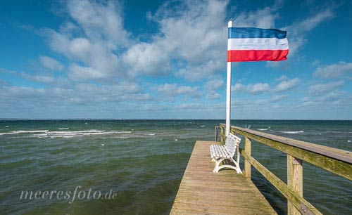 Eine Bank und eine Schleswig-Holstein-Flagge im Sturm auf einem Steg am Strand von Niendorf