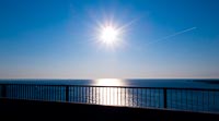  Blick in die Sonne von der Fehmarnsundbrücke –  DETAIL: Die Sonne steht tief über dem Horizont der Ostsee. Das Ende eines Tages auf der Sonneninsel Fehmarn. Im Vordergrund sieht man das Geländer der Fehmarnsundbrücke, ein Flugzeug zieht seine Bahn in nord-östliche Richtung über die »Vogelfluglinie«.