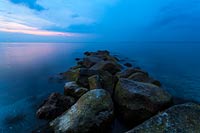  Steinbuhne am Strand von Heiligenhafen, mit Blick auf den Leuchtturm Flügge –  DETAIL: Kräftiges Blau färbt den Himmel zu dieser späten Abendstunde. Das Foto zeigt eine mit Moos und Algen bewachsene Buhne. Am Horizont erkennt man den Leuchtturm von Flügge und einen Teil der Insel Fehmarn.