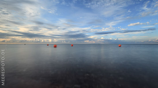  Bojen zur Markierung von Bootsliegeplätzen vor Hohenfeld –  DETAIL: Bei leichter Meeresdünung bewegen sich die großen orangenen Bojen sanft auf der Wasseroberfläche und verschwimmen so auf diesem Langzeitbelichtungs-Foto. Die Sonne geht bald unter und am Horizont sammeln sich die Wolken zur Nacht.