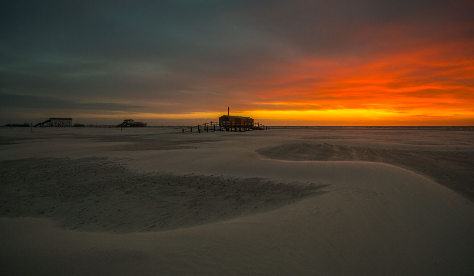Ein Strandschuppen aus Holz am Strand von St. Peter-Ording mit sehr farbintensiven Abendrot nach einem sommerlichen Sonnenuntergang.